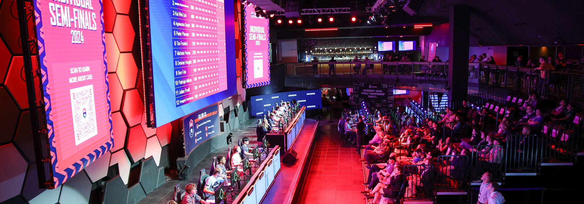 The MECC championship arena in Las Vegas, competitors at their stations along a lit stage with leaderboards on the big screens and an audience watching.