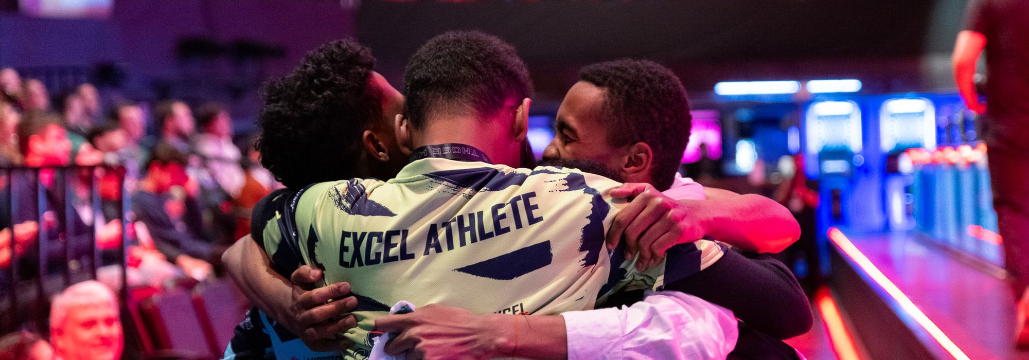 Three competitors embracing in celebration on the competition floor, one wearing a jersey that reads Excel Athlete.