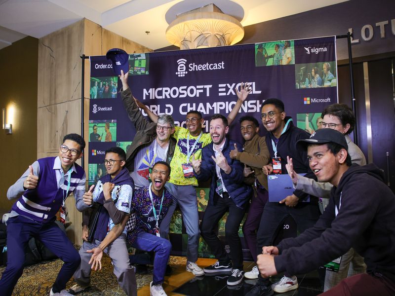A group of competitors posing energetically in front of a Microsoft Excel World Championship backdrop.